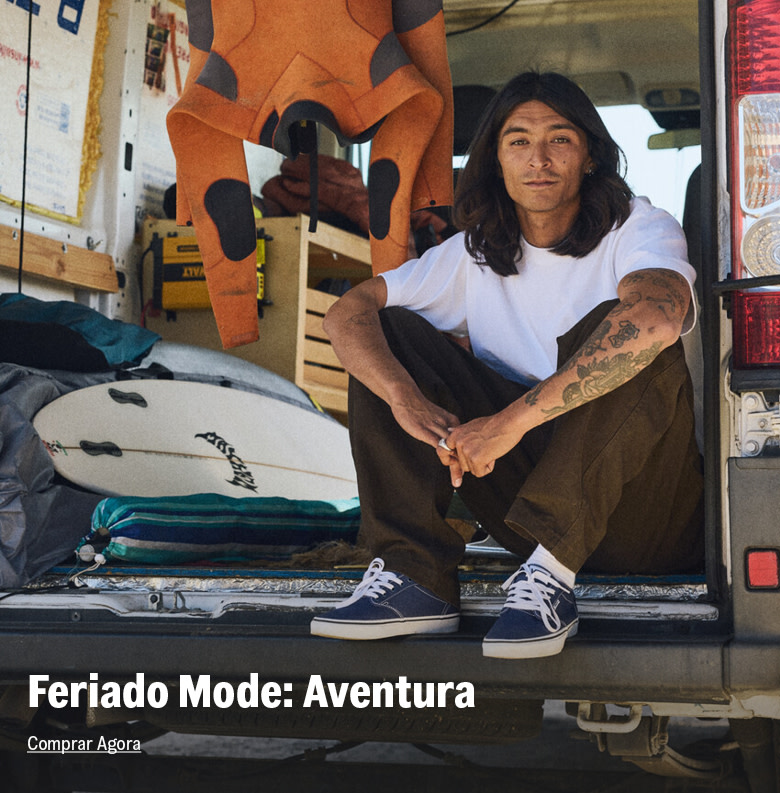 Homem sentado na traseira de uma van com equipamentos de viagem e surf, usando tênis de aventura.