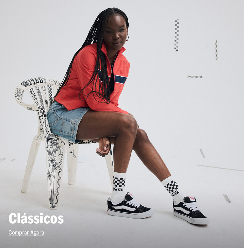 Modelo feminina usando jaqueta vermelha, shorts jeans e tênis Vans Knu Skool preto e branco, sentada em um ambiente branco.